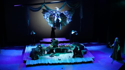 Gliko, in a fantastical feathered costume, descends over five feasting, terrified sailors on a dramatic blue-lit stage.