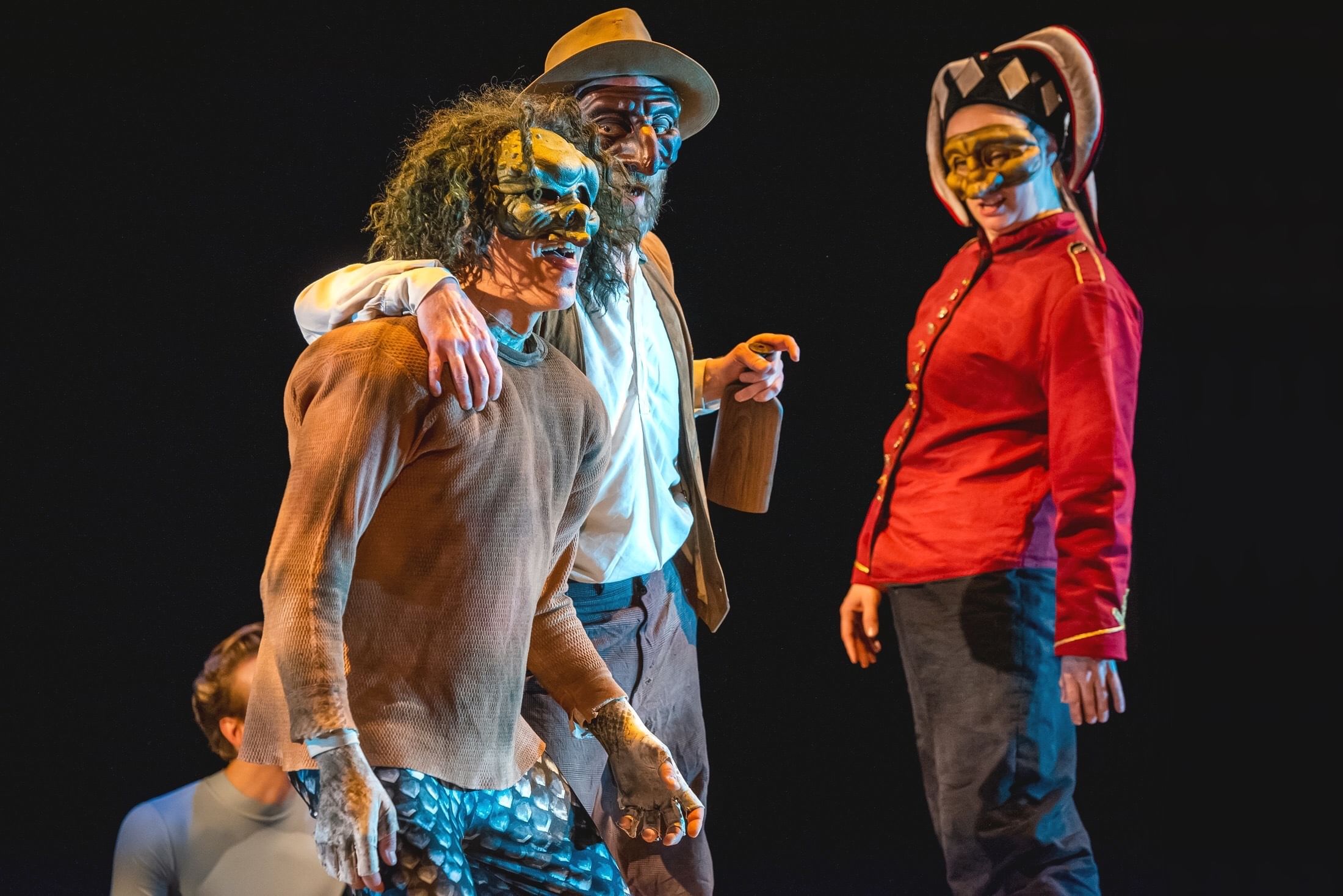 Three actors conspiring in fanciful, somewhat creepy masks, two with hats and one with a wild gray curly wig.