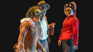 Three actors conspiring in fanciful, somewhat creepy masks, two with hats and one with a wild gray curly wig.