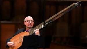 Stone plays a theorbo, a string instrument with a long neck and strings. Stone is on stage, with a music stand in front