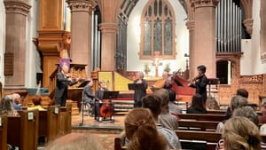 View from the audience in the church pews of six musicians playing period instruments on the chancel.