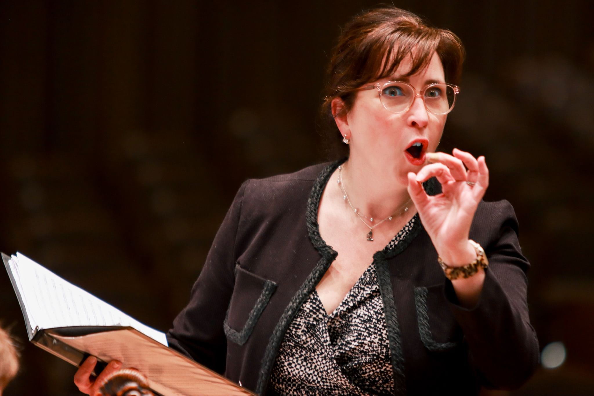 Close-up on Bragle, a white woman with brown hair and glasses, singing animatedly from a handheld score in a binder.