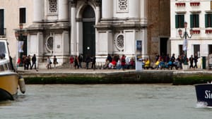 Exterior view of the tall, white, pillared, historic, tile-roofed Venice church on a canal. People fill the sidewalk in front