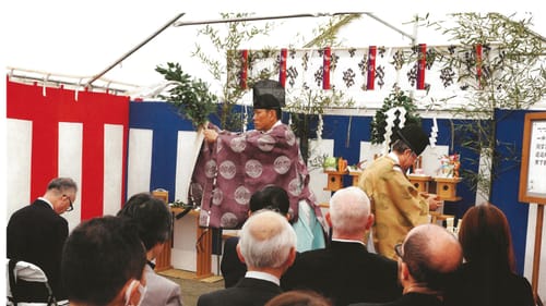 In a white, blue & red tent in front of a seated audience, two men in traditional Japanese garb perform a ceremony.