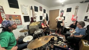 Five people work together in a crowded music studio full of amps, laptops, foot pedals, and instruments.