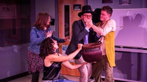 The four actors, in a classroom, strike hectic poses around a woven basket, one covering another’s mouth with his hand.