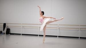 Golz poses in arabest in white tutu. In a rehearsal area with barre bars along the white wall, daylight seeps in from windows