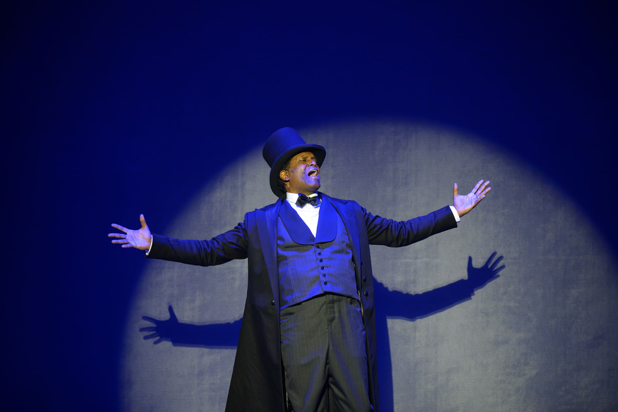 Smiling, a Black man wearing a 19th-century suit and top hat, sings and stretches out his arms in a bluish spotlight.