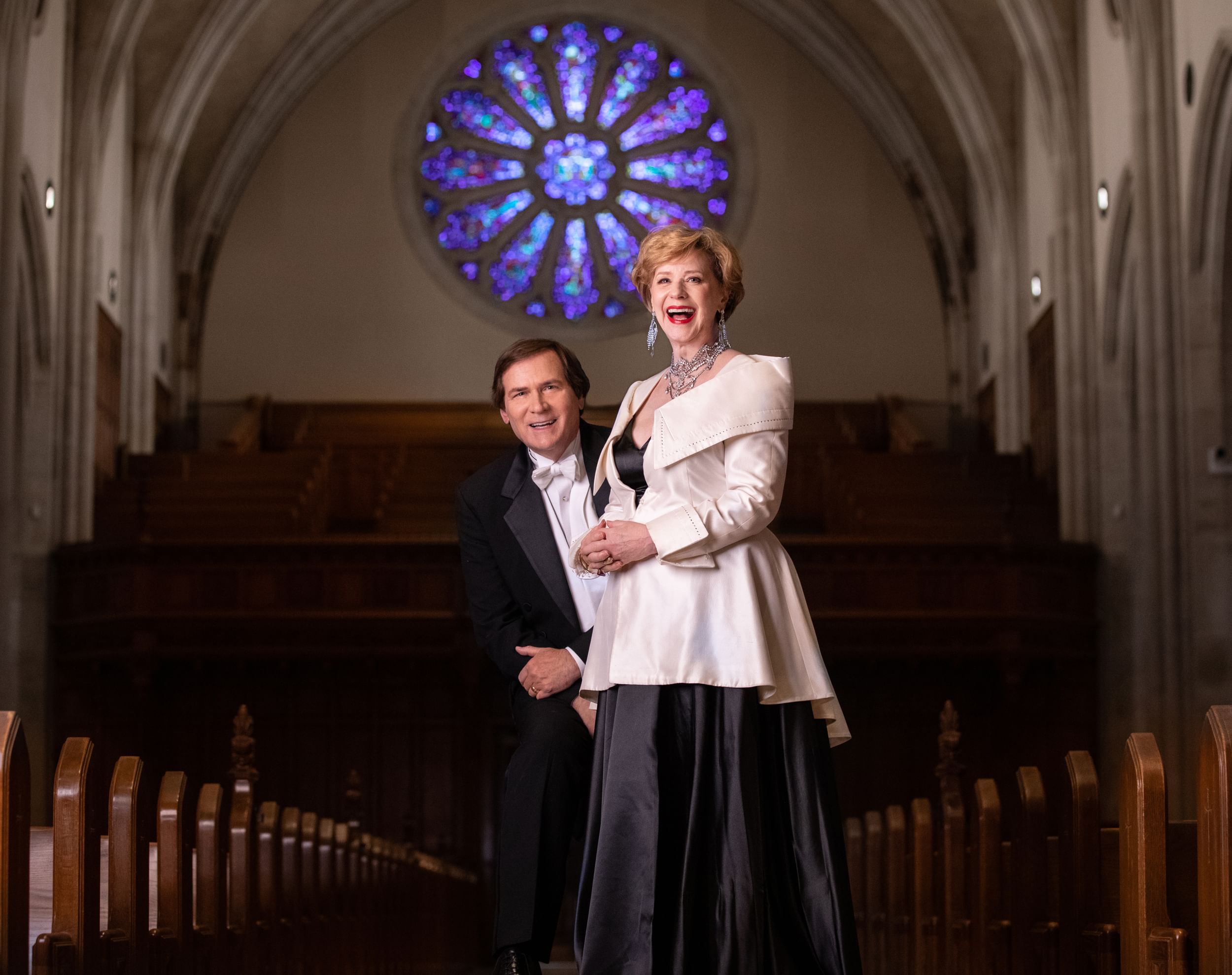 Two people in formal wear stand together, smiling wide for camera, in an empty church with rows of pews behind them