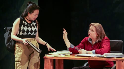 Park, in a plaid sweater and khaki cargos, stands arguing with Rosentrater, wearing a red blouse and seated at a messy desk.