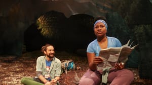 Fayle, in hiking clothes, sits rubbing his foot and smiling at Gardner, who sits on a stump looking annoyed and holding a map