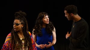 Bowers, a Black woman in a colorful sweater & eyepatch looks away as Gliko and Bellow argue behind her on a bare stage