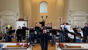 Nally, facing a choir of 16 standing on a church’s white chancel, conducts the singers in a rehearsal.