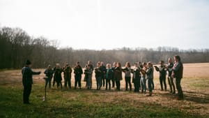 An ensemble rehearses outside in a wide open field under an overcast sky, a conductor stands before them with a podium.