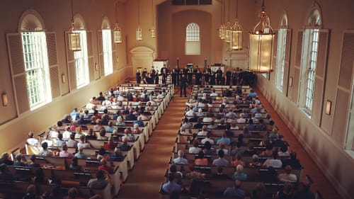 An aerial view from the upper rear of a church, people crowded in all of the pews listening to a choir sing up front.