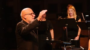 A close-up on conductor Donald Nally, a bald white man, during a choir performance. He is smiling and wears a dark suitcoat.