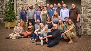 25 choir members, men and women posing together on an old brick patio in casual clothes outdoors, next to a stone building.