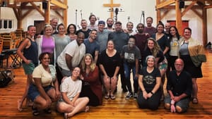 A multi-racial group of about two dozen people posing together in a simple church venue with high wooden rafters.
