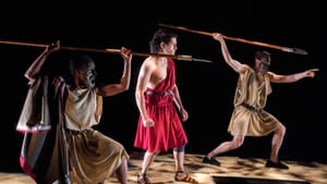 A scene from the show. Ramirez at center wears a red toga, and two chorus members pose on either side with long spears.