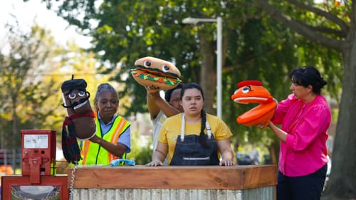 Four actors hold a total of three large hand puppets: an éclair, a banh mi, and a macaron. They’re performing in a park.