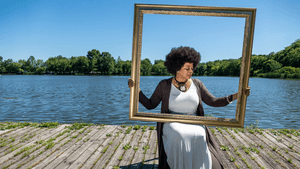 Floyd, a Black woman, sits on a river dock on a sunny day, holding a gold frame in front of her. She wears a long pale dress