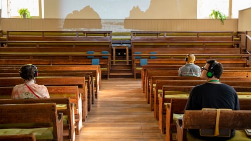 Three people sit with their backs to the camera in the early 1800s wooden pews of the meeting house facing a video projection