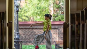 A woman dancer in a green shirt and loose striped pants poses in the doorway of the meeting house, a brick wall behind her.