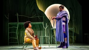 Fabiyi, a Black actor with an afro, wearing earthy yellow clothes, sits on a stool and speaks to Lally, who wears a blue robe