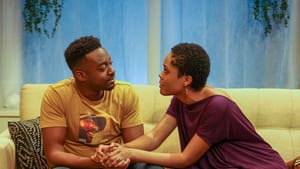 Sesay and Bey, two Black actors, speak intently to each other while sitting on a yellow couch, their hands clasped together.