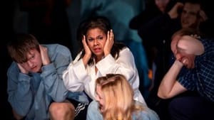 A person centered, dressed in white, holds their hands up to their ears in disgust, others are around her doing the same