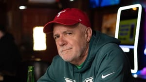 Graham, a white man with white stubble, wearing an Eagles jersey & Phillies hat, sits in a bar holding a beer bottle.