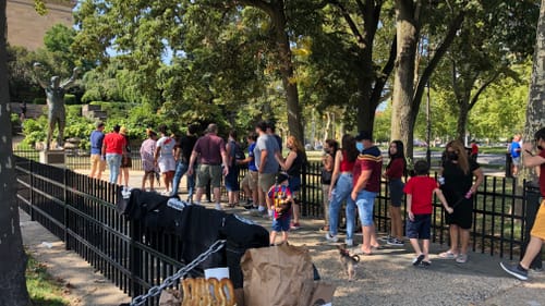 About 20 people stand in line to visit the Rocky Statue outside the museum. Pretzels, drinks, & shirts are for sale in front
