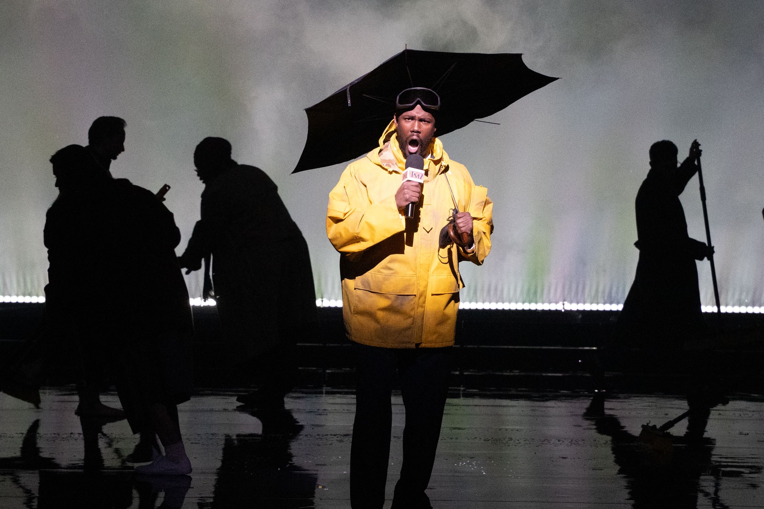 Mburu sings dramatically into a newscaster’s mic, wearing a battered yellow slicker, as silhouetted people dodge around him.