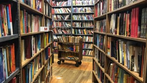 A photo from between two high bookshelves in The Strand, facing more bookshelves, and a rolling cart full of books.