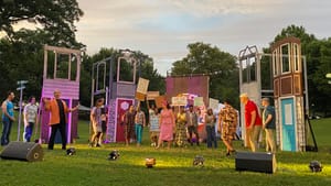 Dusk falls on a scene from the play depicting a protest with the whole cast on the green lawn. Set pieces evoke rowhouses.