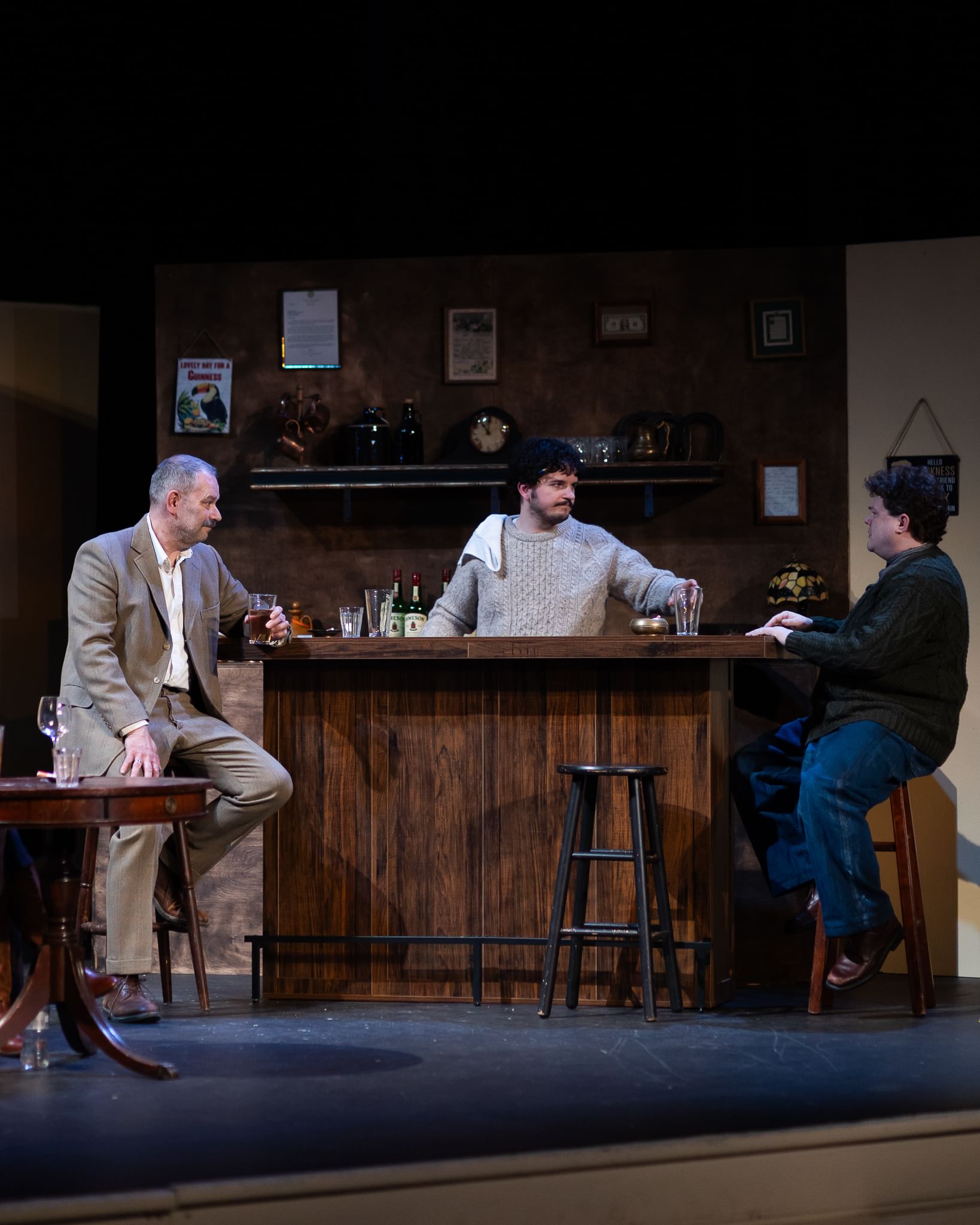 McDonald, in a pale sweater, tends bar at center. Hargraves, in a gray suit, and Donahue, in dark sweater, sit at either end.