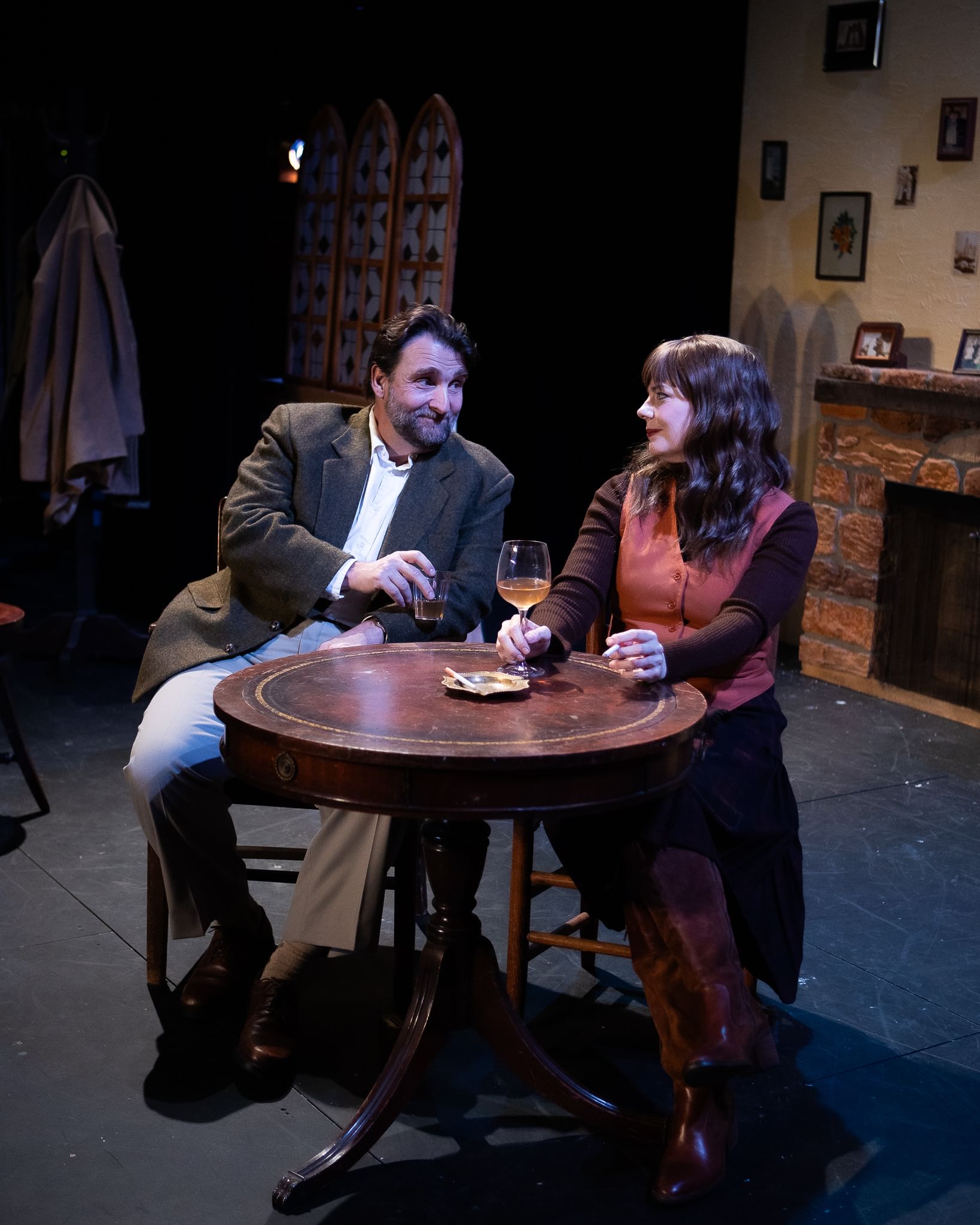 Rock sits at a small table with Quinn, smiling and leaning toward her with a shot glass. She looks wary, holding a wineglass.