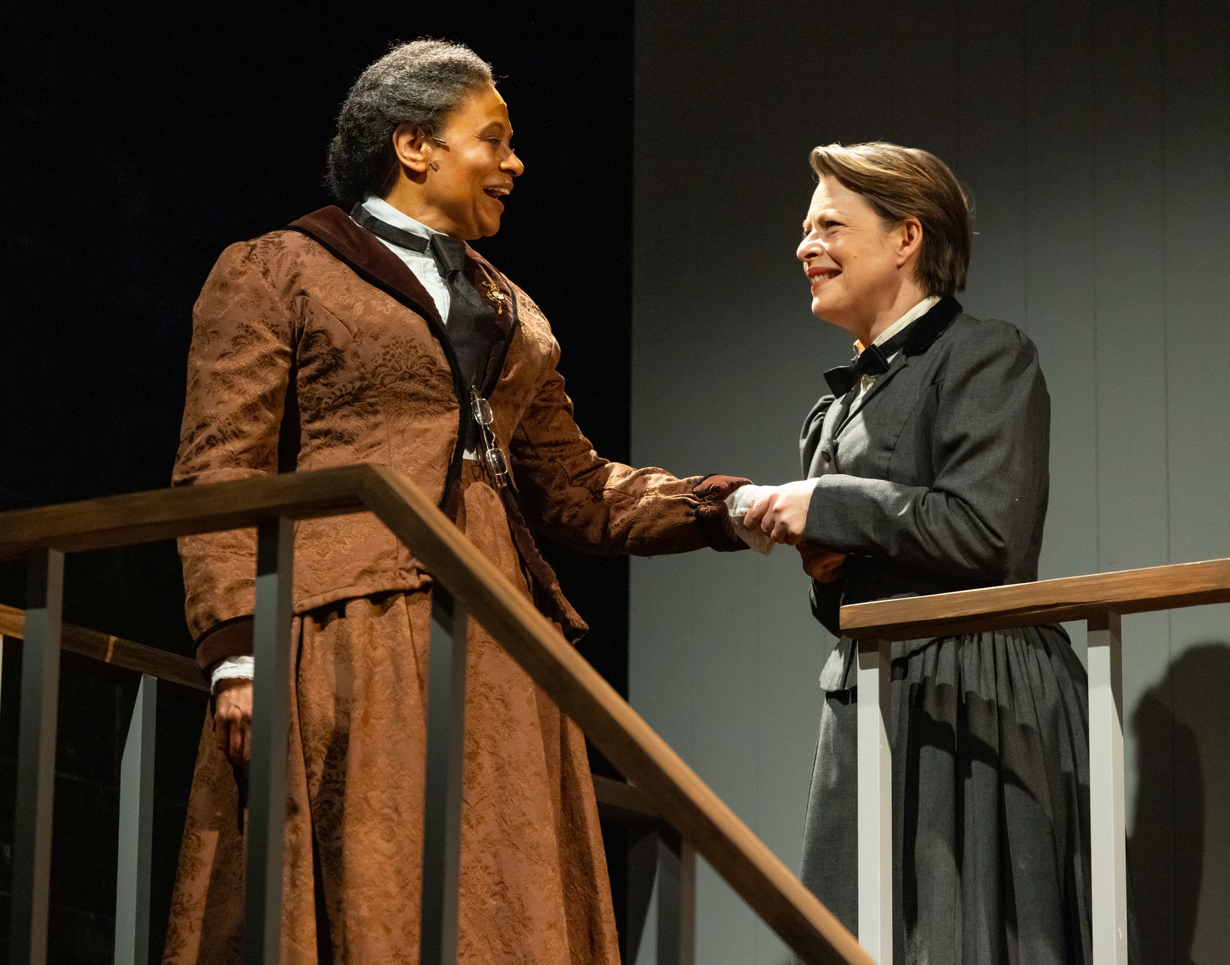 Finister & Holum, in late 19th-century professional garb, smile and clasp hands at the top of a staircase on the set.