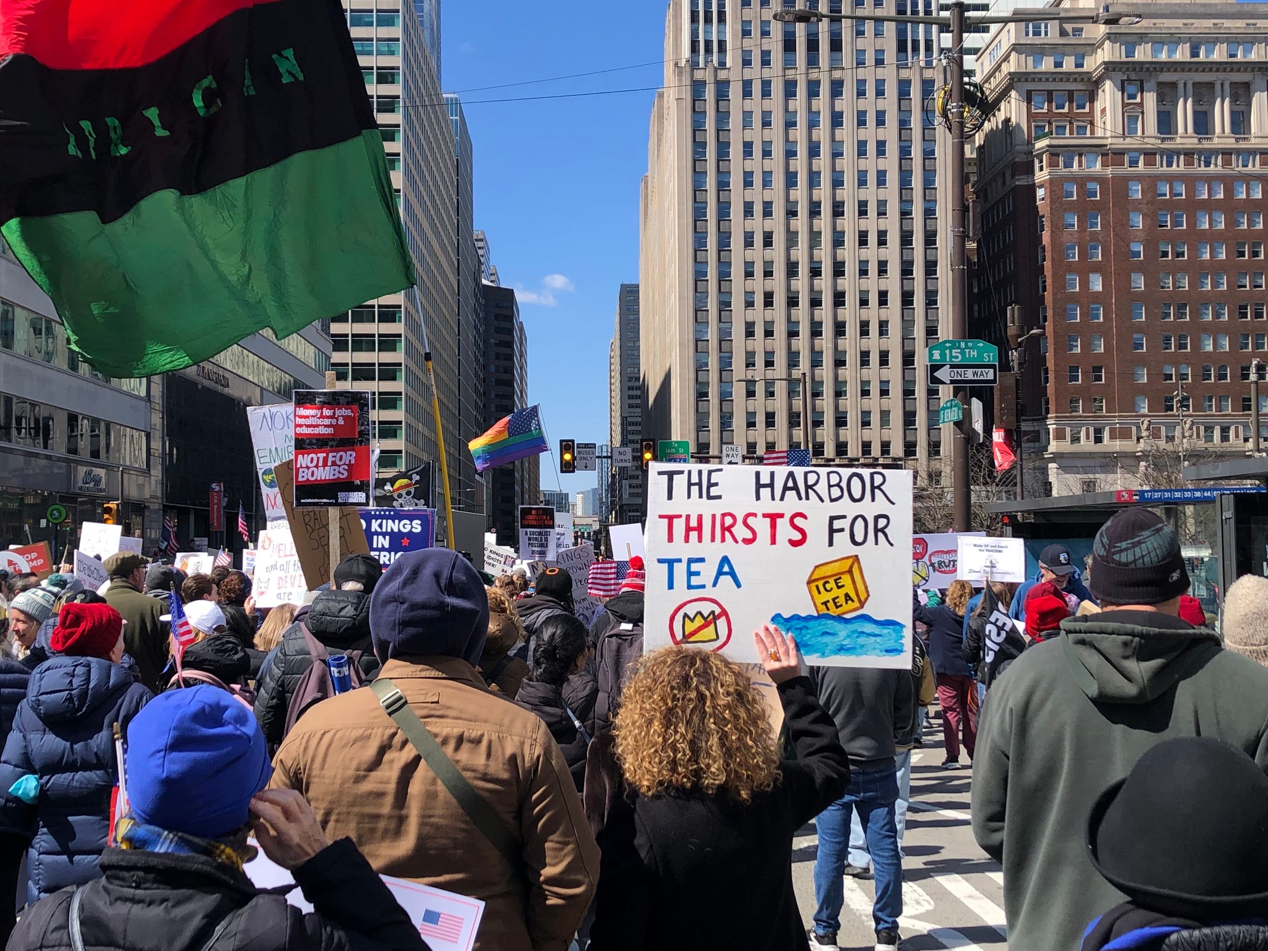 View from inside protest on a sunny, cold day showing hundreds of people from behind, many holding handmade signs.