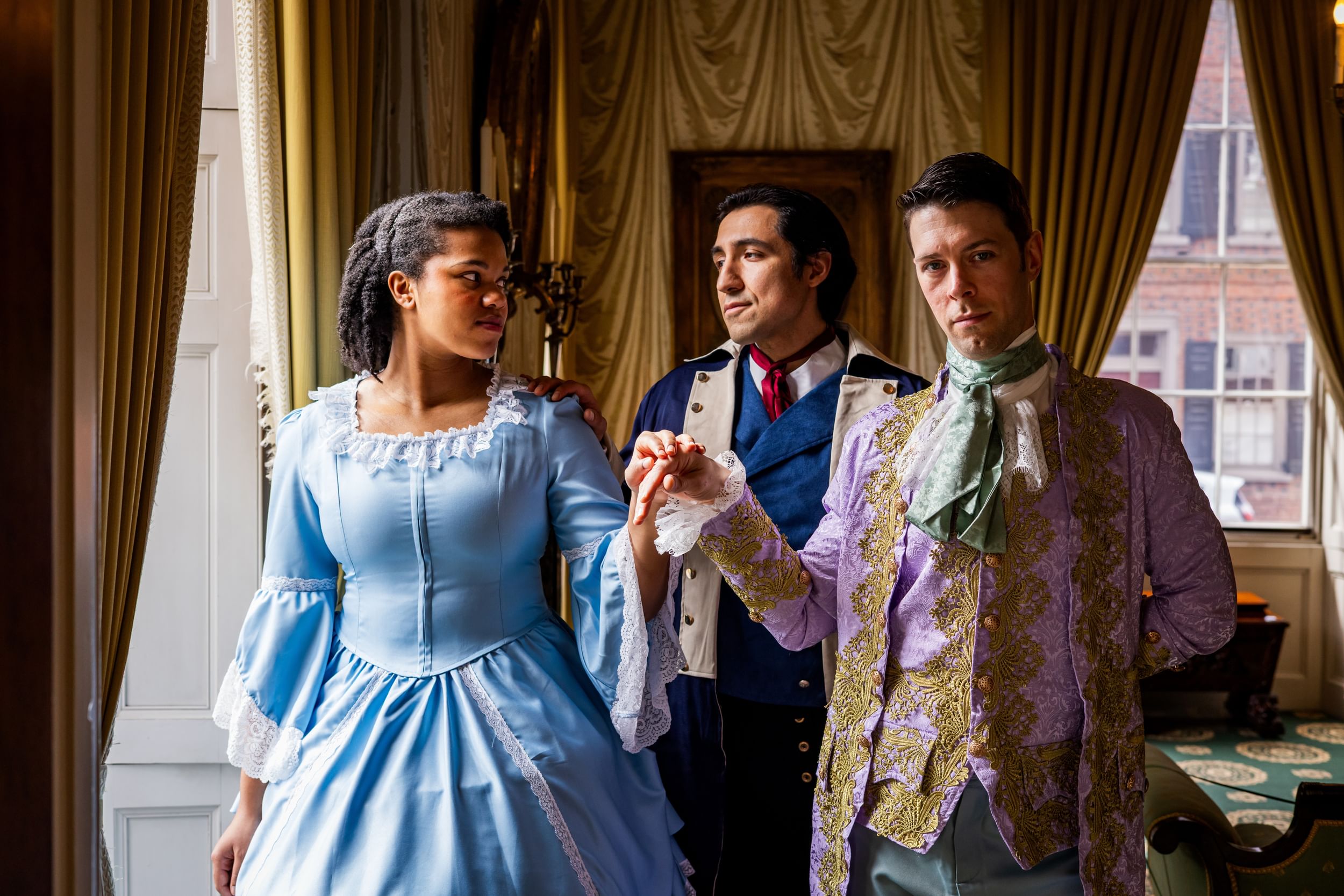 Three people in colonial outfits stand by a window. Two in front hold hands, one of them looking at the other