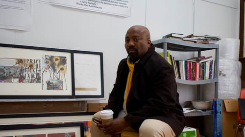 Harris, a Black man in khaki pants, black shirt & yellow scarf, sits on a folding chair next to a bookshelf & framed collages