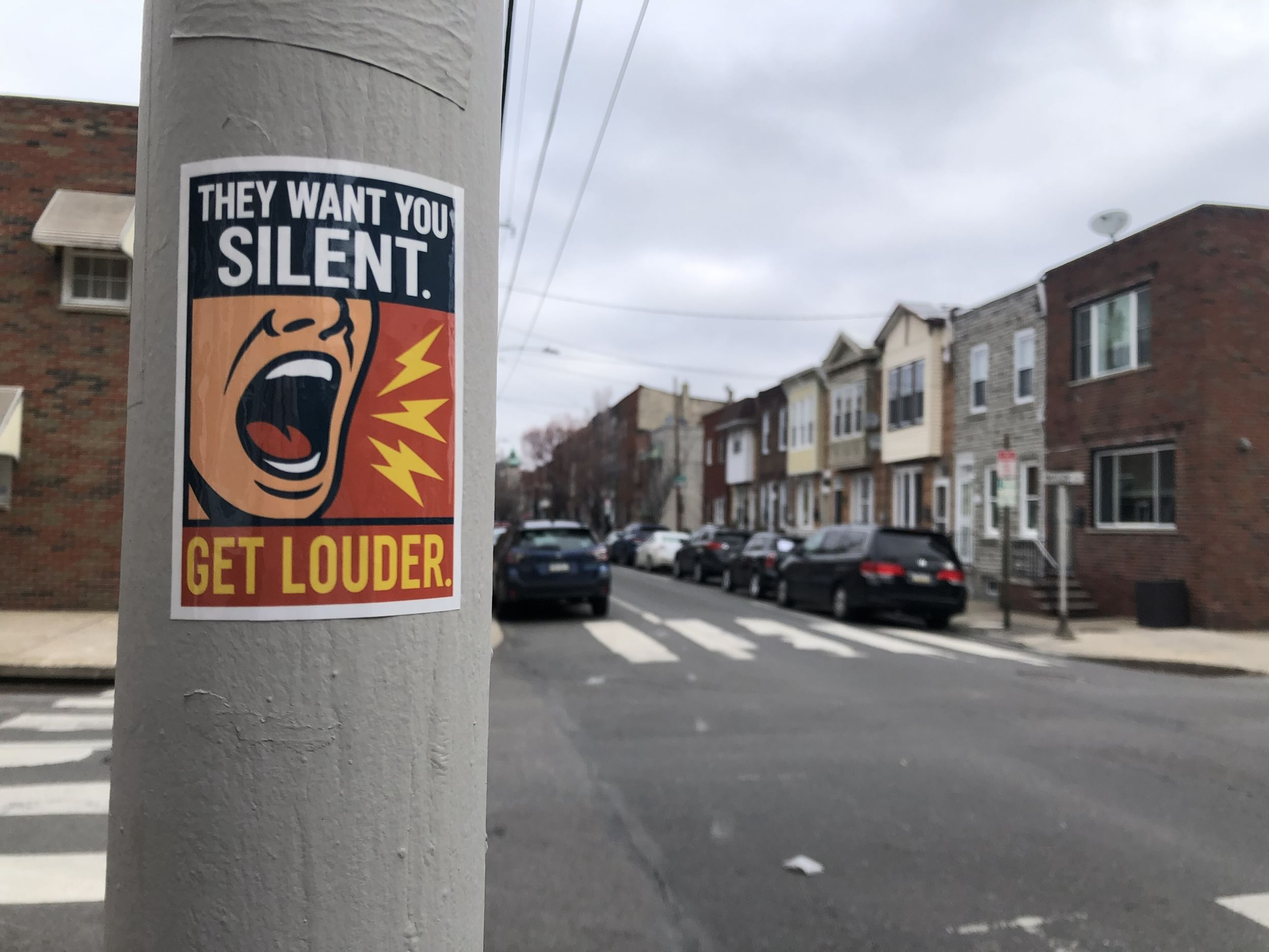 Sticker on a city telephone pole says "They want you silent. Get louder," with an illustration of a shouting mouth.