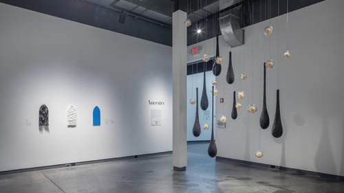 Gallery view. At left, 3 small works on a white wall. At right, Lee’s hanging work of glass balls & elongated black tar drops