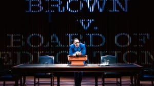 Marable as Thurgood, in a blue suit & glasses, leans on a large table, the words Brown v Board of Education projected behind