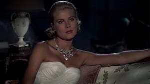 Film still. Actress Grace Kelly sits, a white lamp off behind her in a dim room, straight-faced, looks off-center from camera