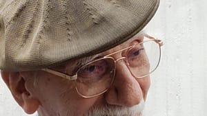 Close-up of Tom, an elderly white man, with glasses, a tan cap, white goatee, and calm smile.