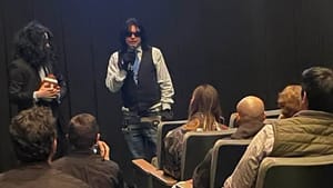 Wiseau wears sunglasses, has a mic, talks to an audience in a small theater room, people are visible in the seats