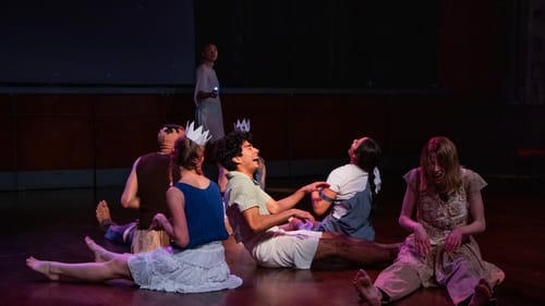 Five dancers in different casual outfits sit on the stage facing different directions, all laughing.