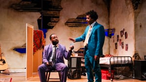 In a dilapidated apartment, St. Pierre wears a purple sit. He sits on a chair & hands money to Merchant, in a turquoise suit