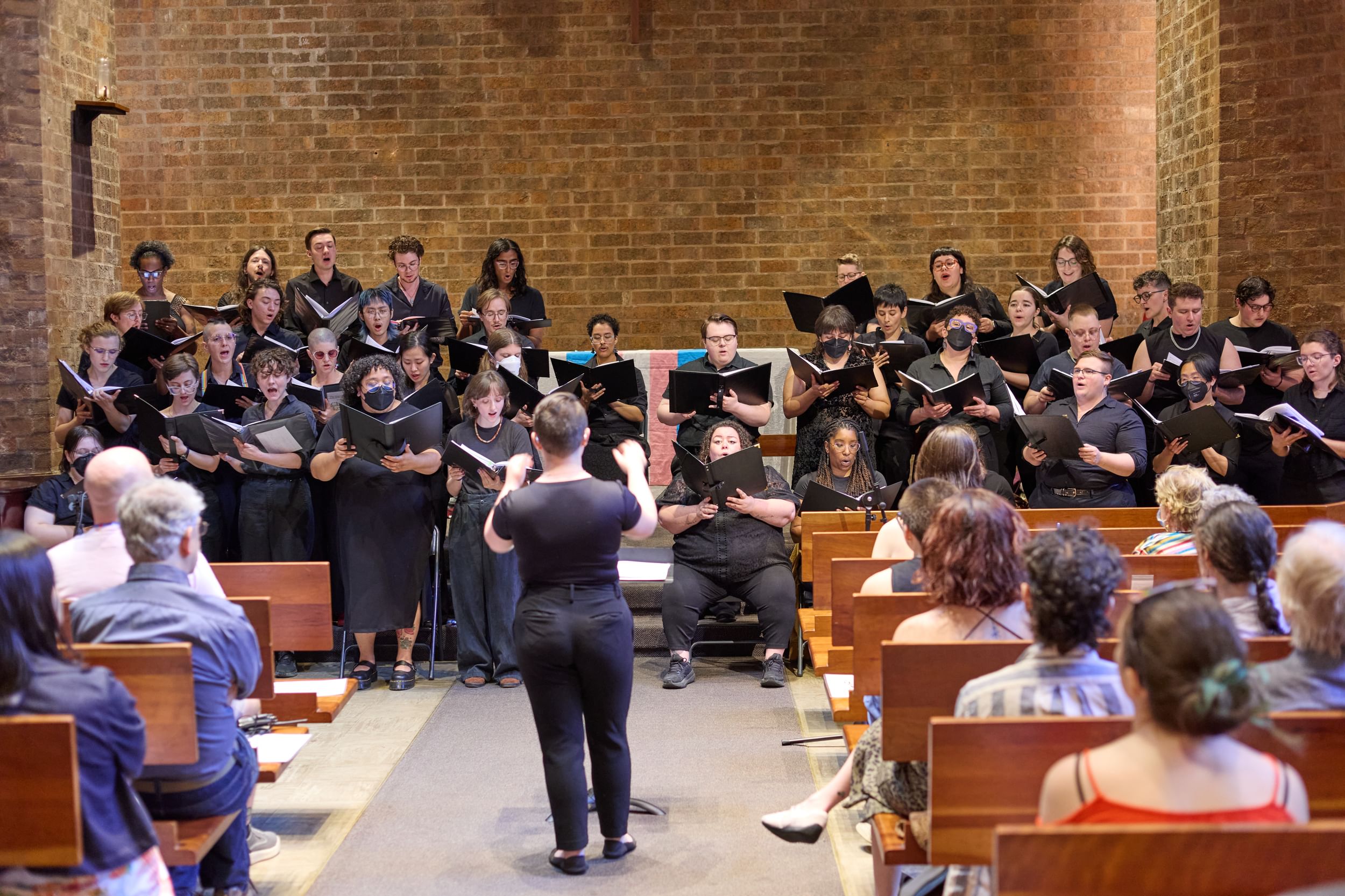 A choir of 35 singers of many genders, all in black, sing against a brick wall backdrop, a conductor in front of them